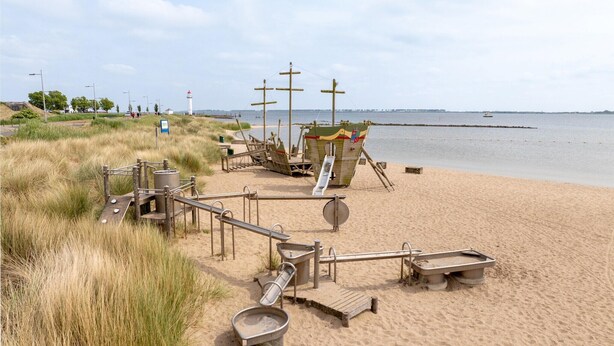 Une plage avec une maquette de bateau en bois, une balançoire et divers conteneurs métalliques.
