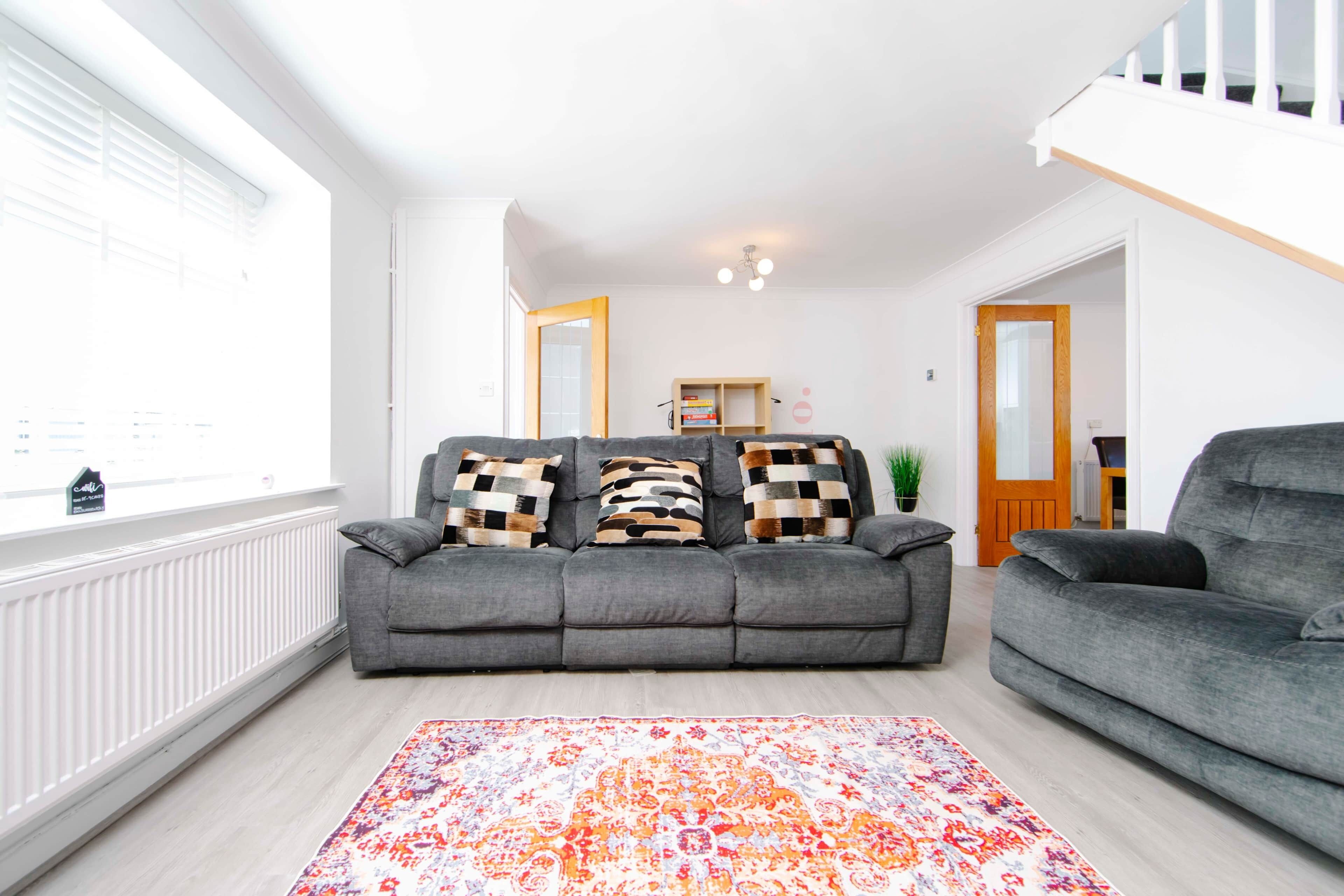 A living room with a grey sofa, patterned cushions, a colorful rug, and a wooden bookshelf.