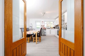A modern kitchen with white cabinets, a dining area with a table and chairs, and wooden doors. - Gilfach Cynon Cottage - Beacons & BikePark (Merthyr Tydfil)