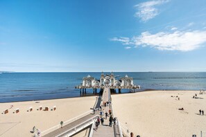 Une jetée menant à un bâtiment à la façade symétrique, une plage de sable et l’océan.
