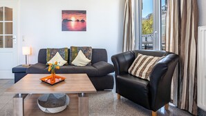 A living room with a sofa, a coffee table with a vase, and a painting of a sunset. - Holiday Apartment 'Am Hochufer - First Row' with Sea View, Balcony and Wi-Fi (Sellin)