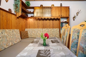 A dining area with a table set for two, a green vase with flowers, wooden paneling, and a bookshelf with books and decorative items.
