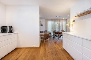 Una cocina moderna con armarios blancos, un comedor con mesa y sillas de madera, y una sala de estar con sofá y cortinas.