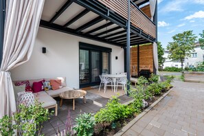 A covered outdoor seating area with a sofa, chairs, and a table, surrounded by plants and a brick pathway.