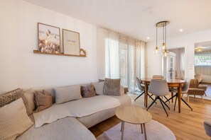 A modern living room with a sofa, dining table, and chairs. There are framed pictures on the wall and a chandelier.
