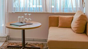 Ein Zimmer mit einer Couch, einem kleinen runden Tisch mit Notizbuch, Gläsern und einem Stift, sowie einem Fenster mit durchscheinenden Vorhängen.