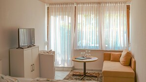 Ein Zimmer mit einem Bett, einem Schlafsofa, einem Fernseher auf einer Kommode, einem kleinen Tisch mit Gläsern und einem Fenster mit leichtem Vorhang.