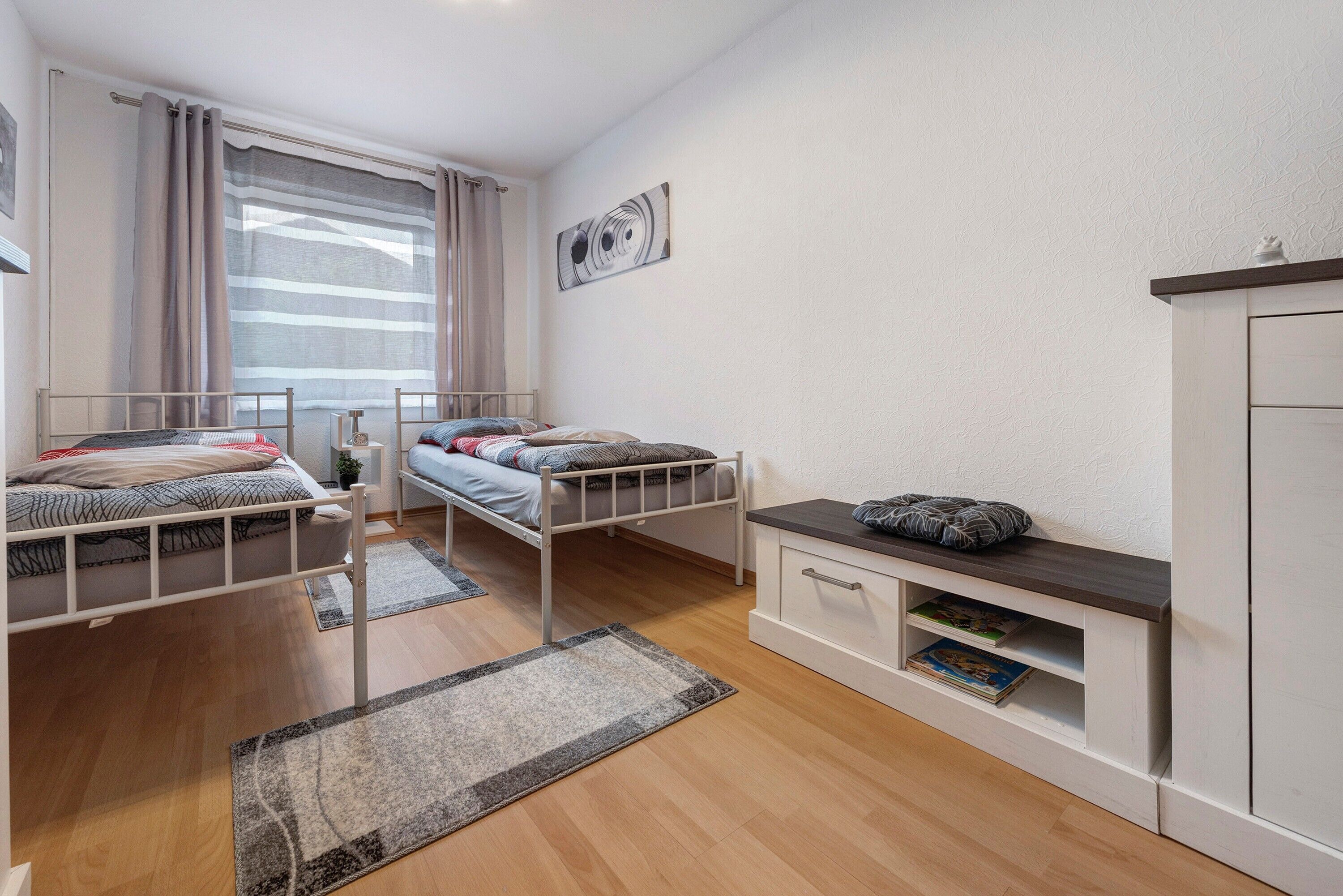 A bedroom with two beds, a window with curtains, a bookshelf, and a wall-mounted artwork.