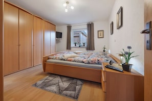 Un dormitorio con cama, armario de madera, televisor, ventana con cortinas y un escritorio con planta.