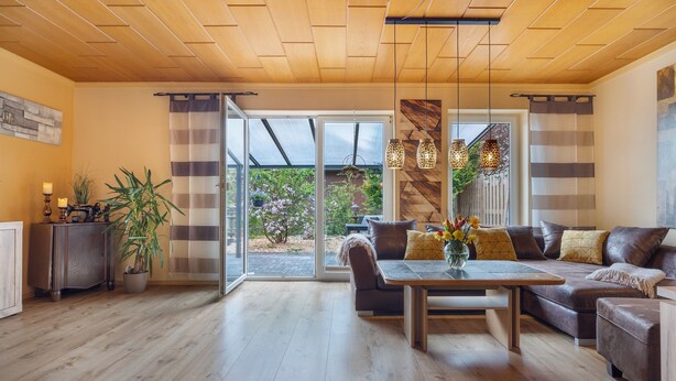 Una sala de estar moderna con techo de madera, un conjunto de sofás, una mesa de comedor y un amplio ventanal corredera que da a un jardín.