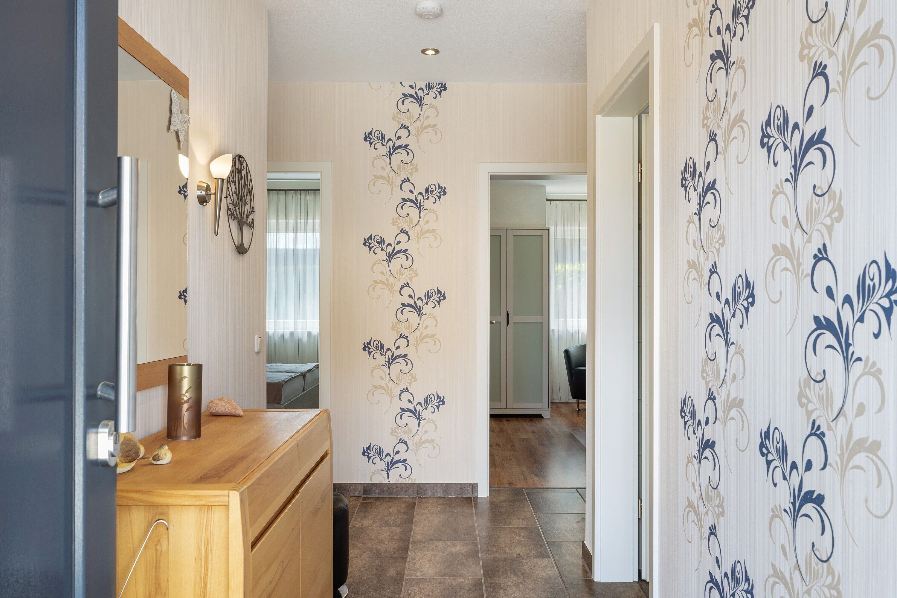 Un couloir avec une commode en bois, un miroir, des appliques murales et un motif décoratif sur le mur.