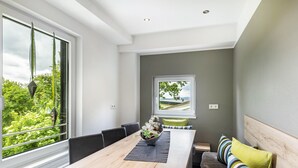 A modern dining area with a wooden table, chairs, a bowl of flowers, and a view of greenery through large windows. - Apartment 'Sonnblick' with Shared Terrace, Shared Garden and Wi-Fi (Schmallenberg)