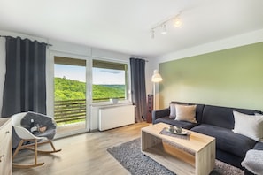 Un salon moderne avec un canapé, un fauteuil à bascule, une table basse et une vue sur un paysage verdoyant grâce à de grandes fenêtres.