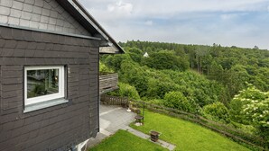 A house with a dark exterior, a window, a paved patio, and a view of a forested hill. - Apartment 'Sonnblick' with Shared Terrace, Shared Garden and Wi-Fi (Schmallenberg)
