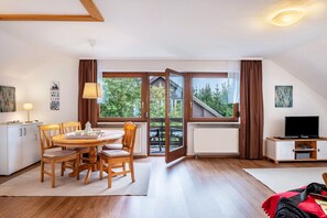A room with a dining table, chairs, a TV, and a view of a house through large windows.