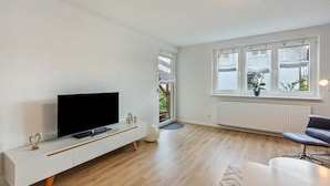 A modern living room with a flat-screen TV on a white console, a large window with a view, and a grey sofa. - Holiday Apartment 'K5' with Private Terrace, Shared Garden and Wi-Fi (Wehr)