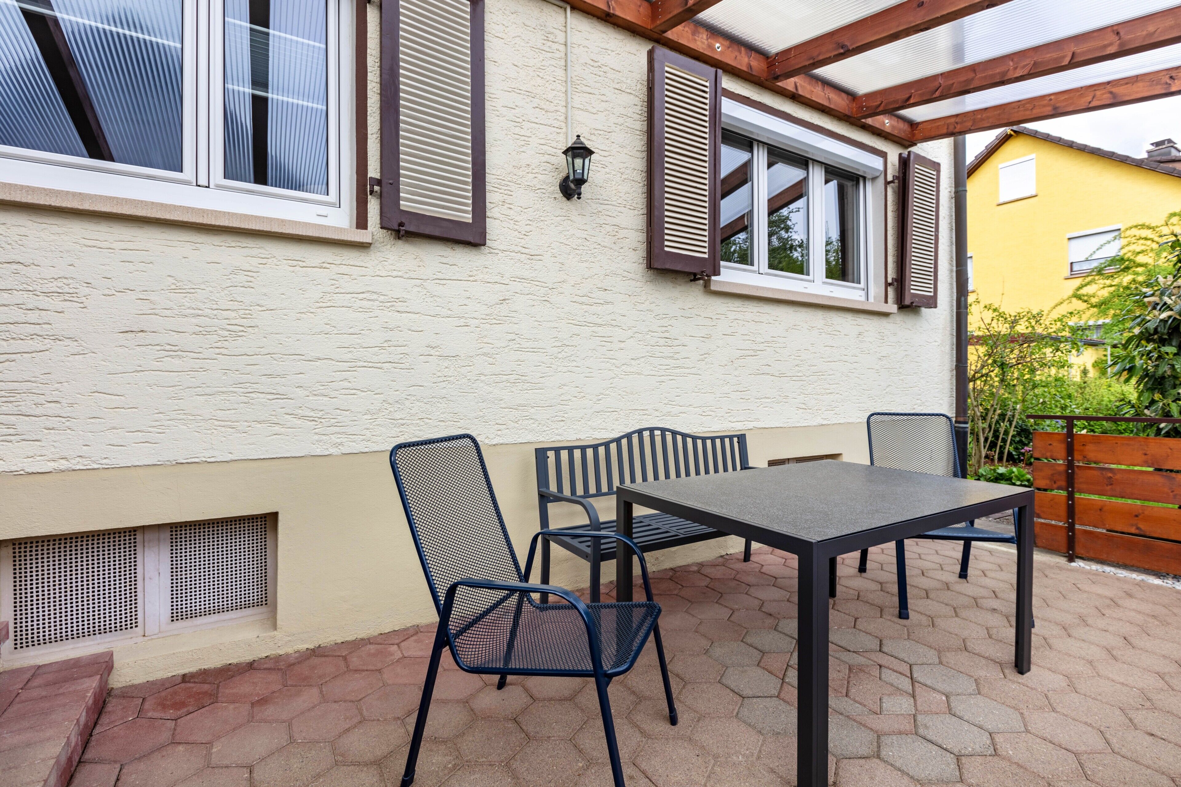 Een overdekt terras met een tafel en stoelen, een raam met luiken en een ventilatie rooster in de muur.