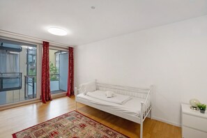 A bedroom with a white bed, a red patterned rug, a small bedside table, and a view through a glass door. - Holiday Apartment 'Aldars 15' with Wi-Fi and Air Conditioning (München)