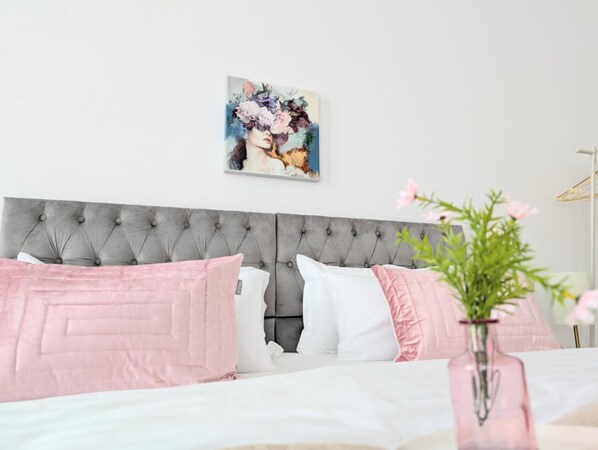 Un dormitorio con cabecera capitonada, cama con ropa de cama rosa y blanca, un jarrón con flores y un cuadro en la pared.