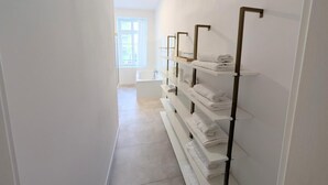 A narrow hallway with white tiled floors and walls, leading to a bathroom with a bathtub and shelves with folded towels.
