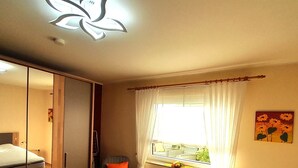 Un dormitorio con cama, armario, silla, ventana con cortinas y un cuadro en la pared.