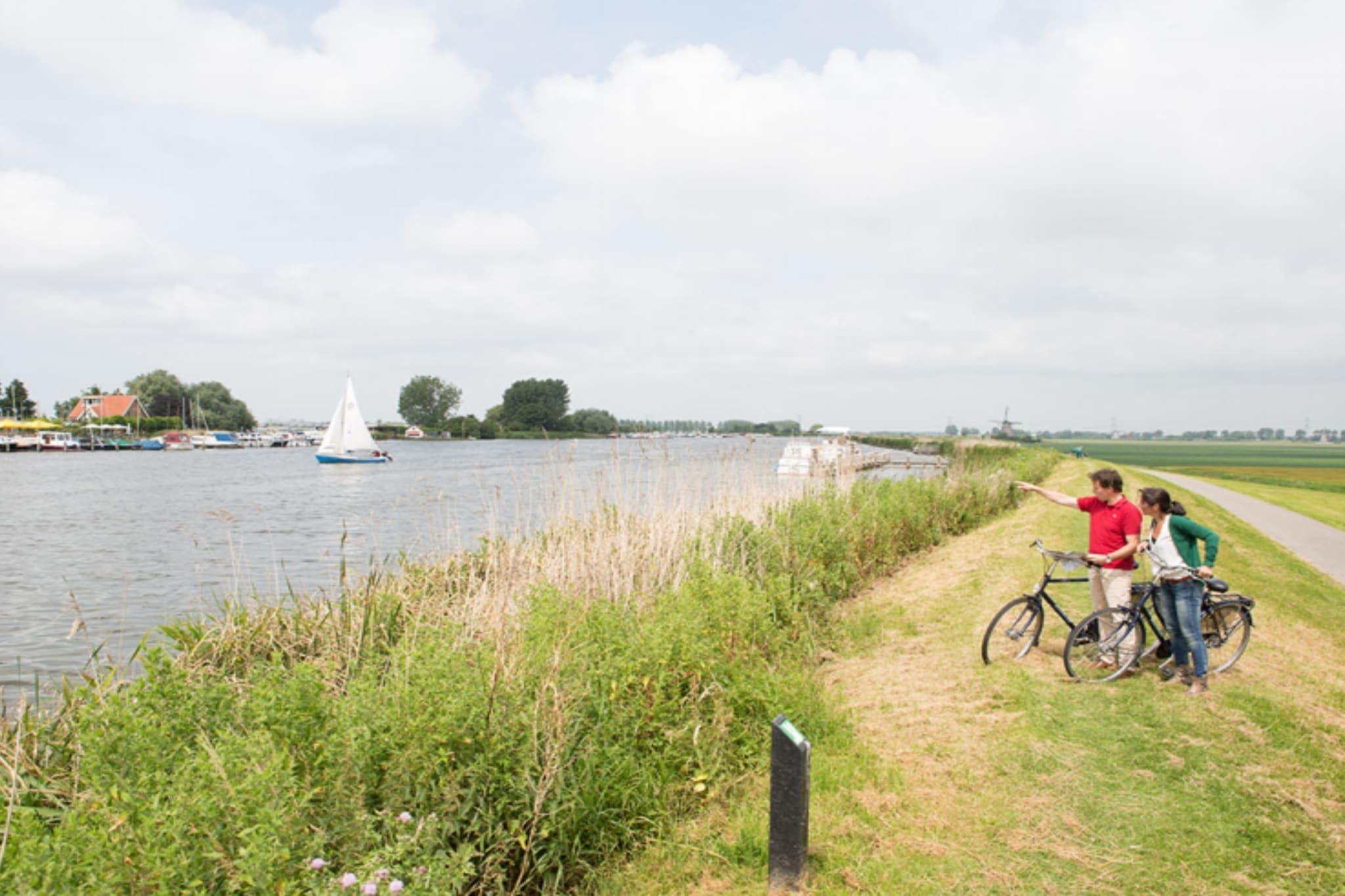 Un sendero junto al río con un velero a lo lejos, un área con césped y una ciclovía.