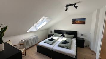 Een slaapkamer met een schuine hemel, een dakraam, een bed met kussens, nachtkastjes, een radiator, een plant en een schilderij aan de muur.