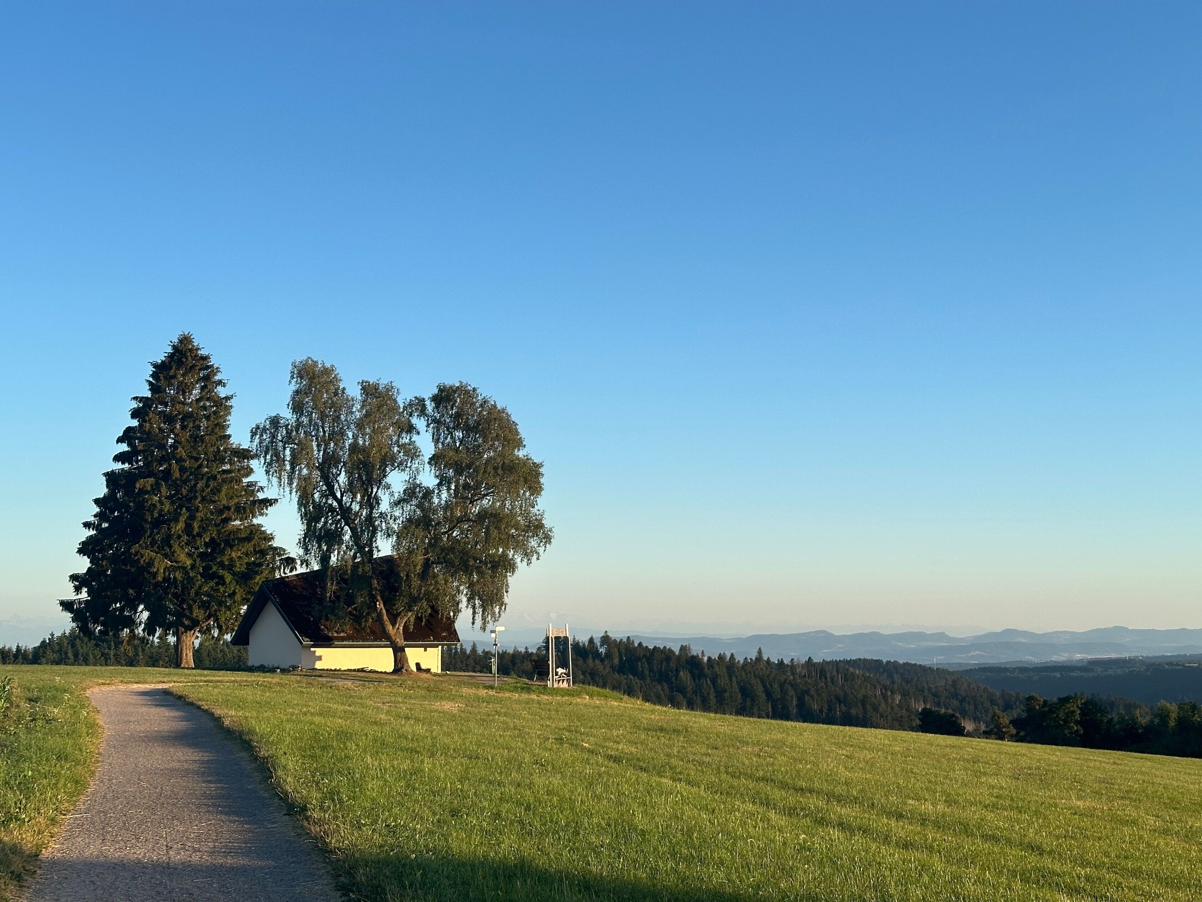 A rural landscape with a path leading to a small building, surrounded by trees and open fields.