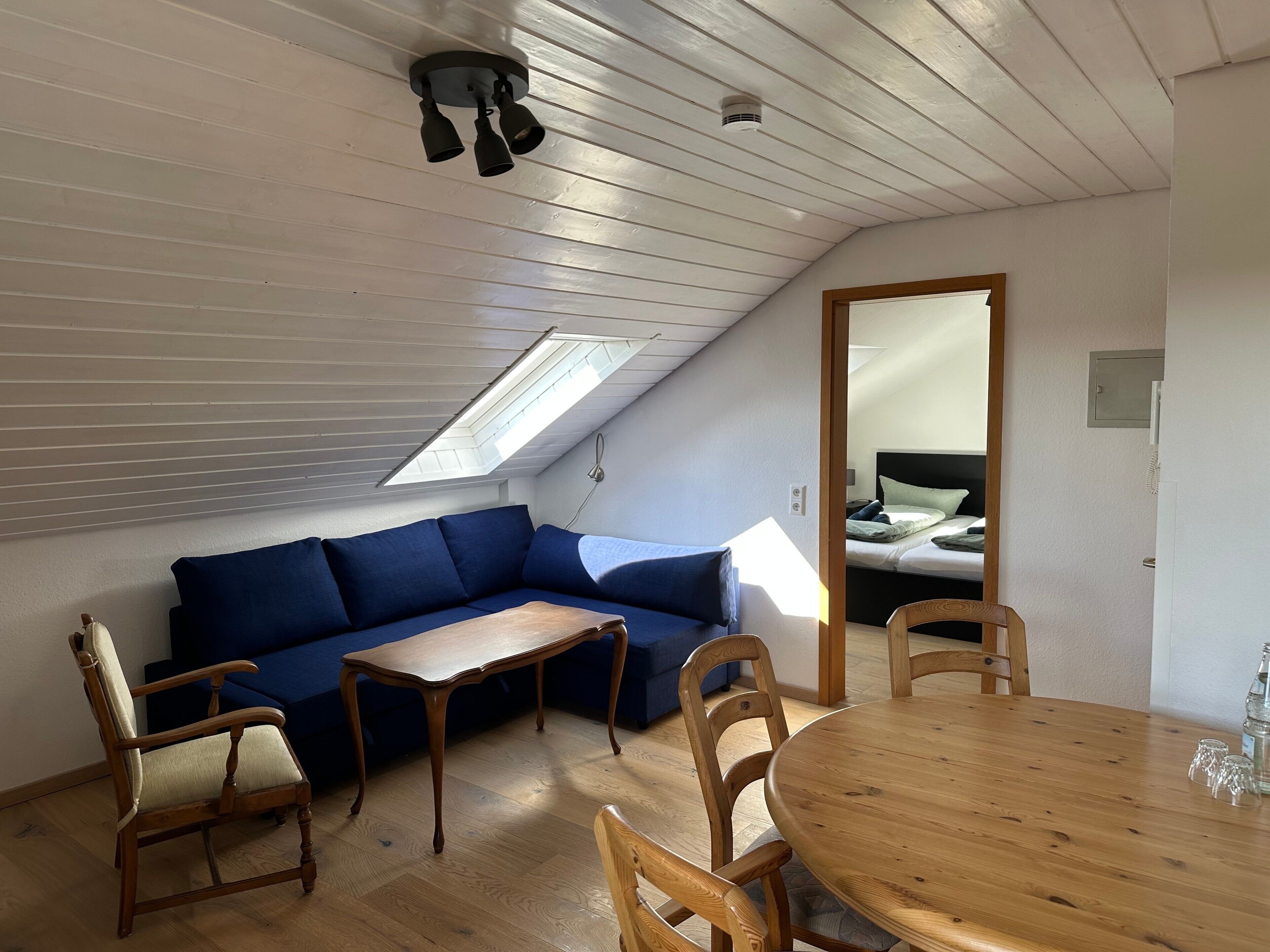A room with a sloped ceiling, a skylight, a blue sofa, a wooden table, and a wooden chair.
