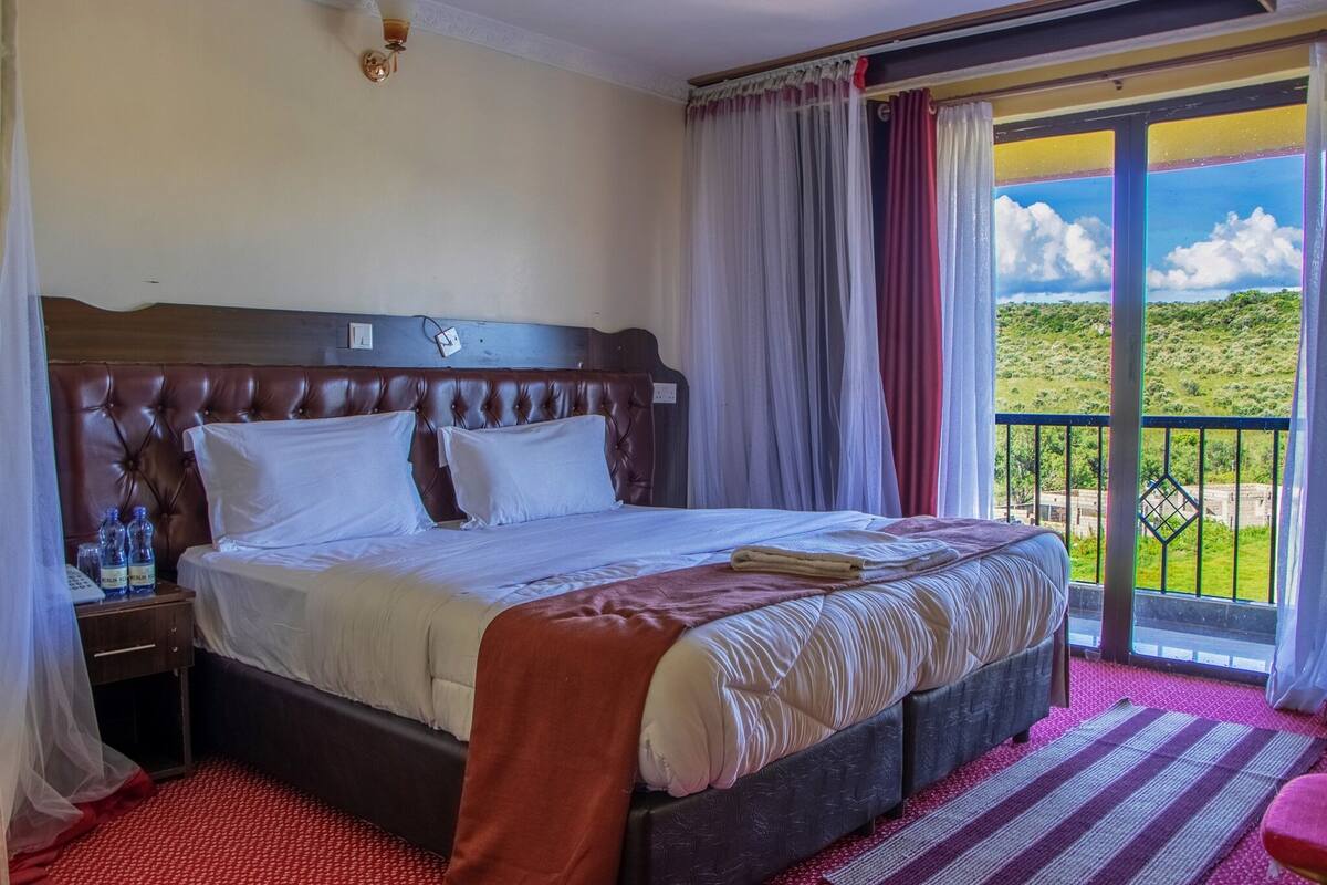 Habitación de hotel con una cama grande, una mesita de noche con un teléfono y botellas de agua, y un balcón con vistas al campo.