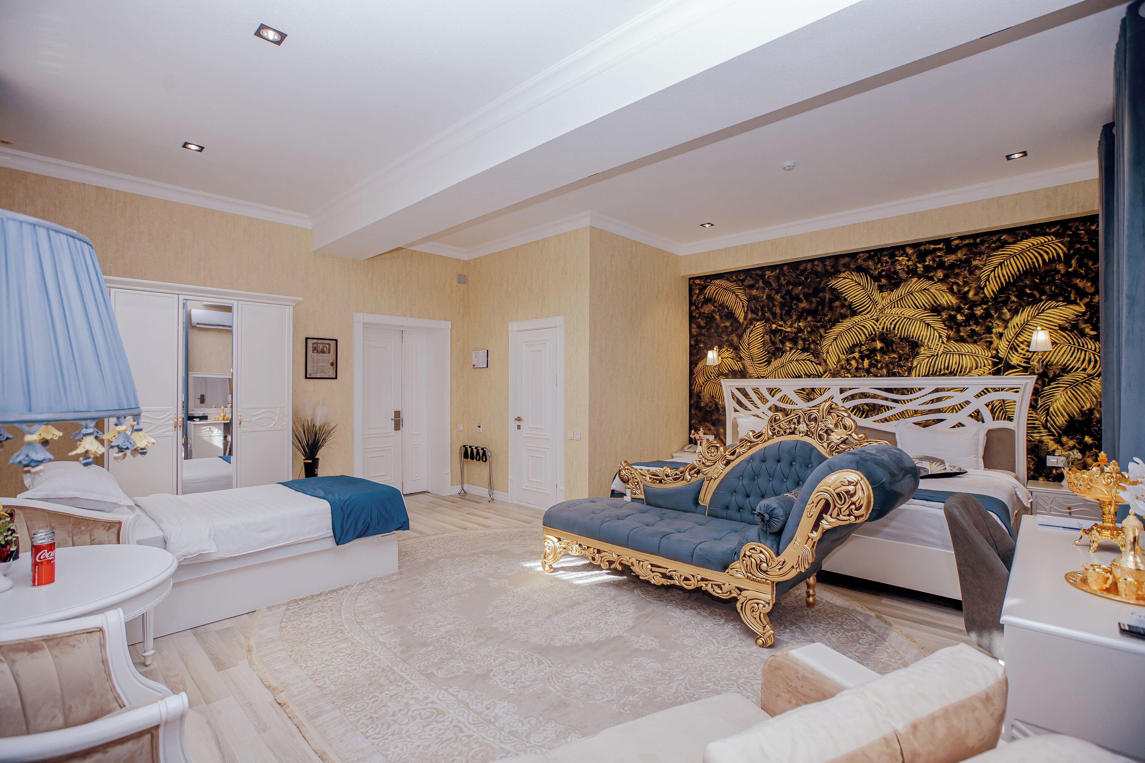A spacious bedroom with a large bed, a sofa, and a desk. There are two doors, a mirror, and a decorative wall panel.