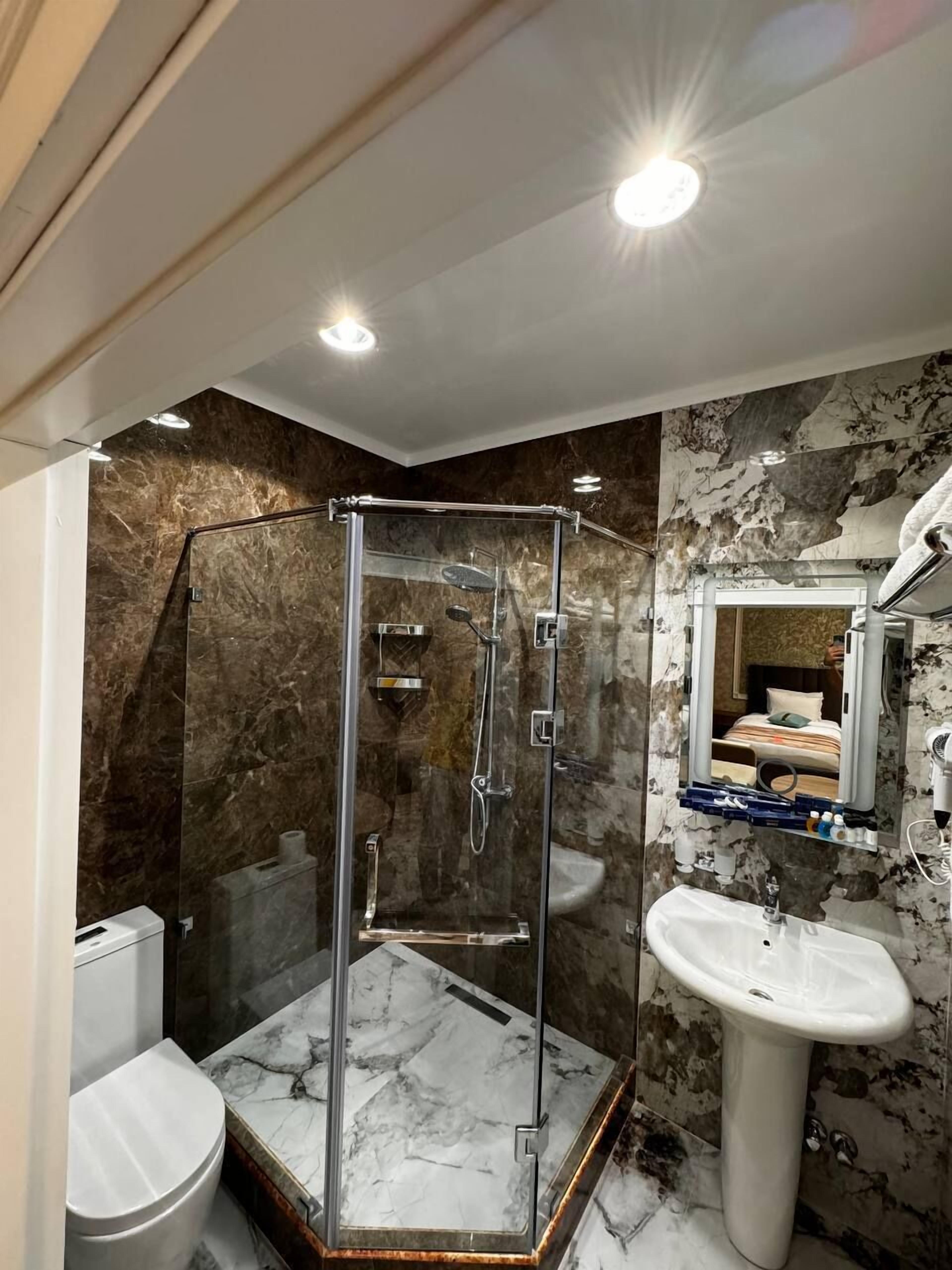 A modern bathroom with a glass-enclosed shower, a marble-tiled floor, a white toilet, and a pedestal sink.