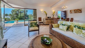 Un espacio habitable amplio con un gran ventanal con vistas a una piscina, un sofá de mimbre con cojines tropicales, un comedor con sillas de mimbre y un mueble con objetos decorativos.