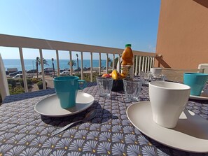 A table set with a bottle of juice, glasses, and plates, overlooking a coastal view with palm trees and a clear sky. - Le Méditerranée by Interhome (Fréjus)