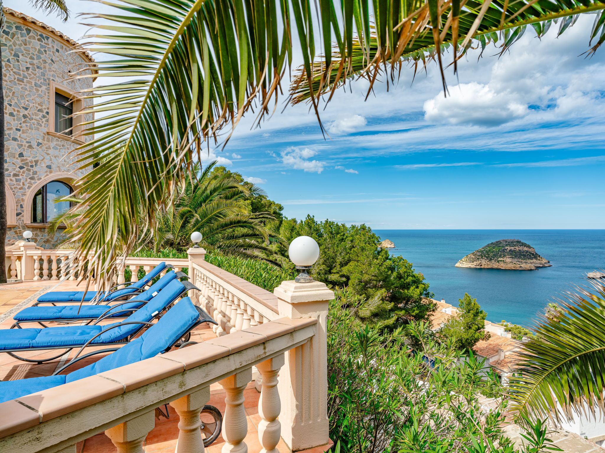 A terrace with blue lounge chairs, a stone building with a balcony, and a view of the sea and a small island.