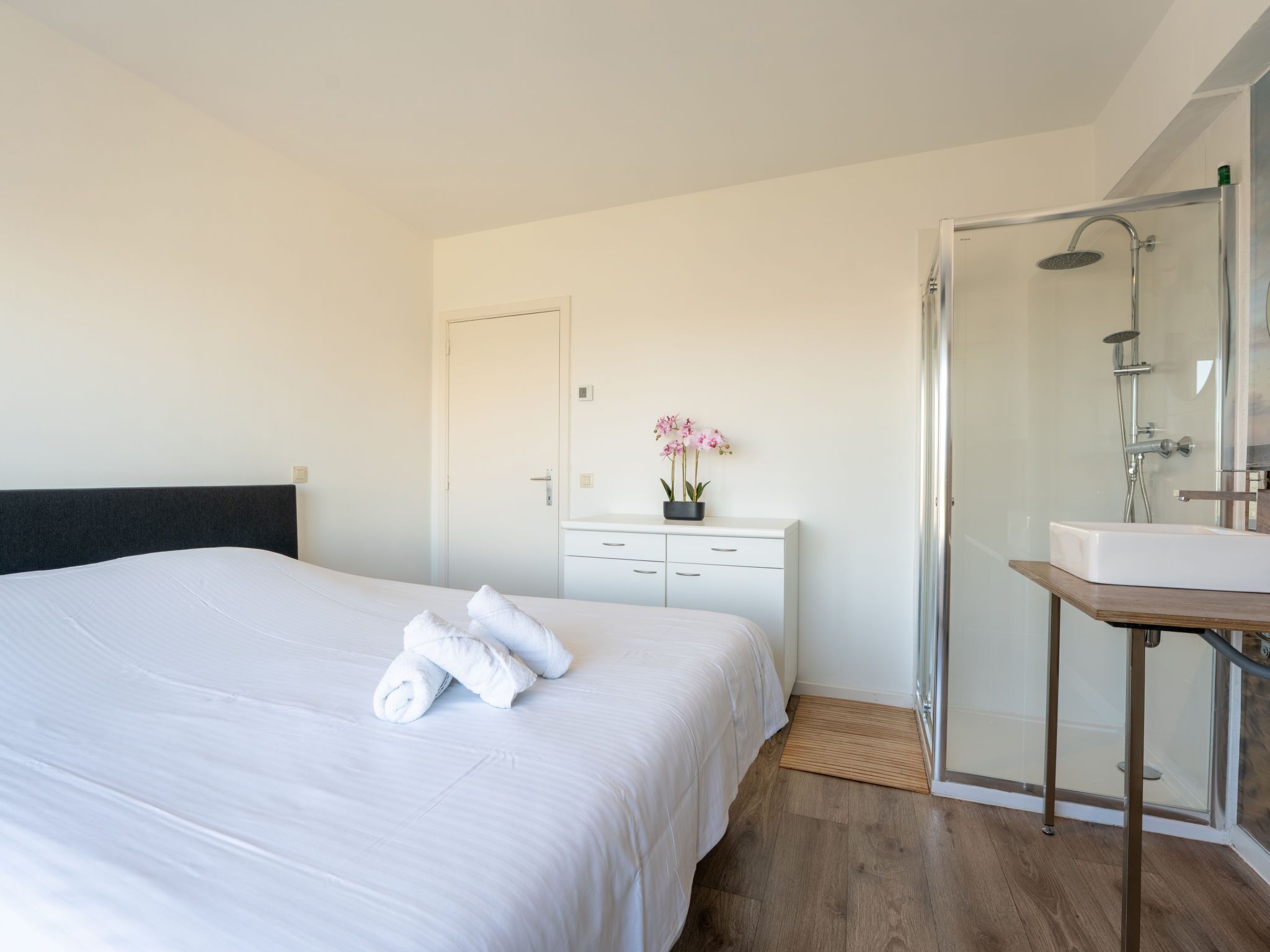 Een hotelkamer met een groot bed, een nachtkastje met een bloevas, een douche en een klein wastafel.