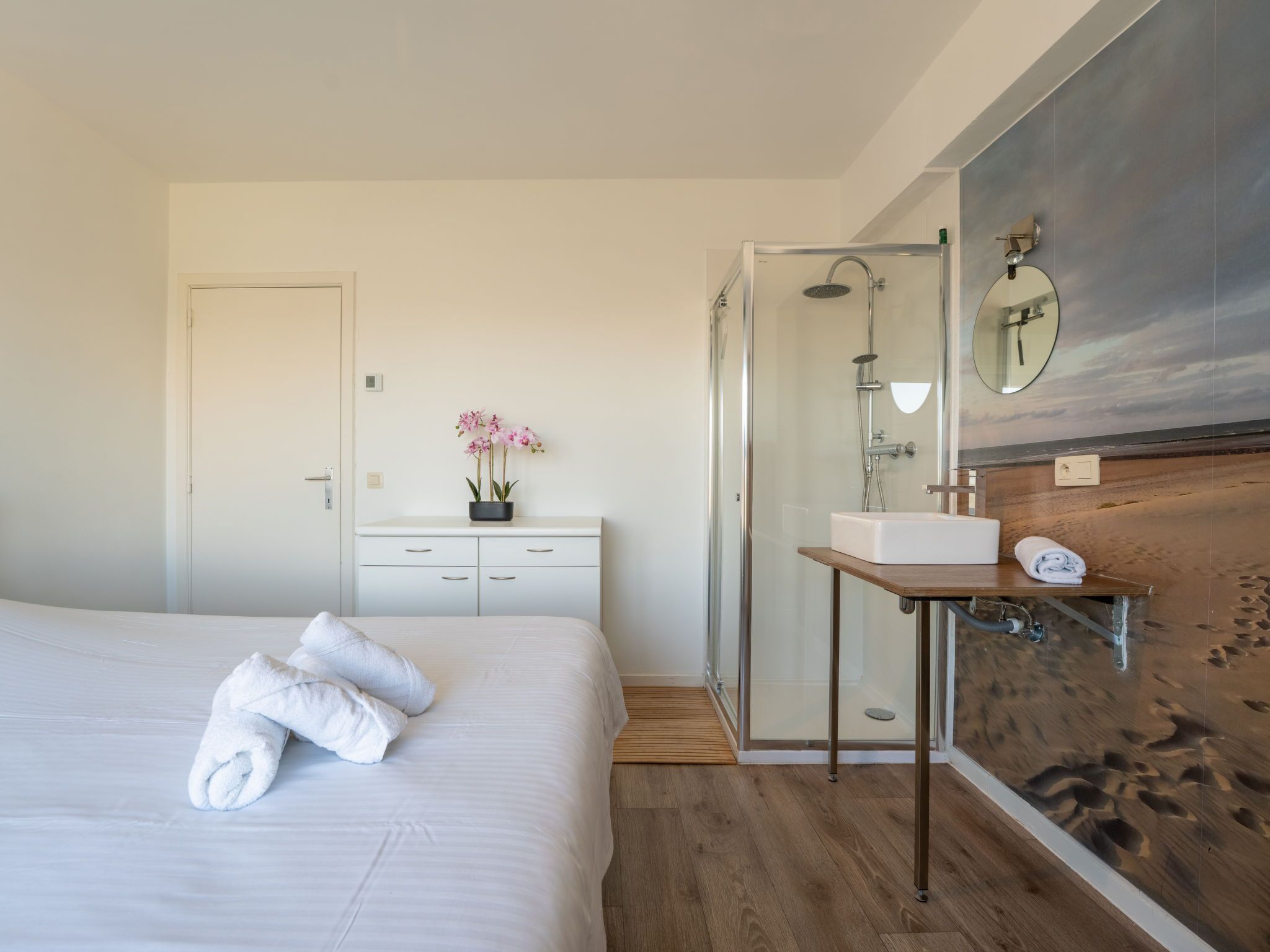 A modern hotel room with a large bed, a bedside table with a mirror, a wooden accent wall, and a glass-enclosed shower area.