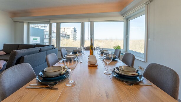 A dining area with a wooden table set for four, large windows, and a view of buildings and grass.