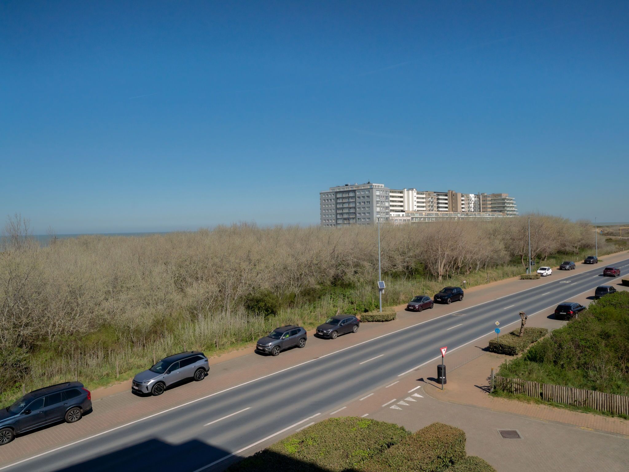 A multi-lane road with parked cars and a pedestrian walkway alongside a grassy area with sparse vegetation.