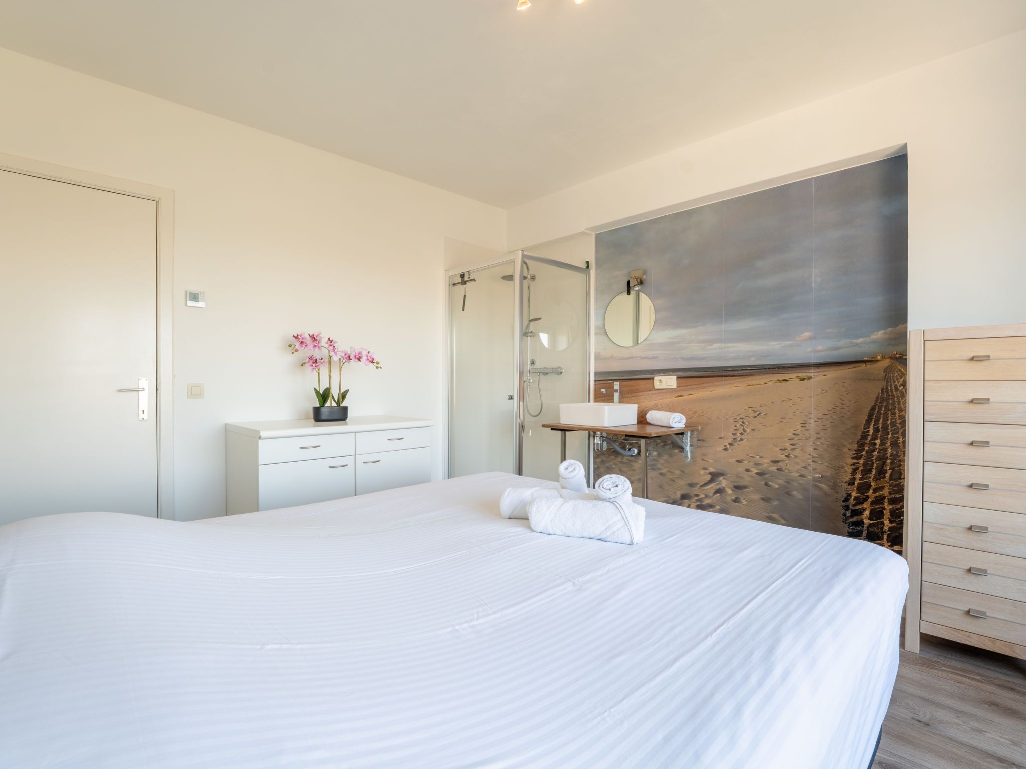 Ein Schlafzimmer mit einem großen Bett, einem Nachttisch mit Orchidee im Topf, einem Badezimmer mit Dusche und Waschbecken sowie einer Wand mit einem Strandgemälde.