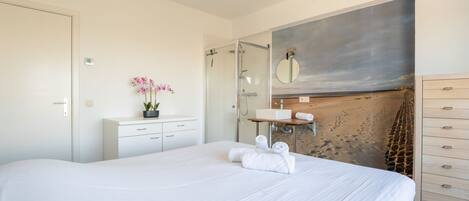 Ein Schlafzimmer mit einem großen Bett, einem Nachttisch mit Orchidee im Topf, einem Badezimmer mit Dusche und Waschbecken sowie einer Wand mit einem Strandgemälde.