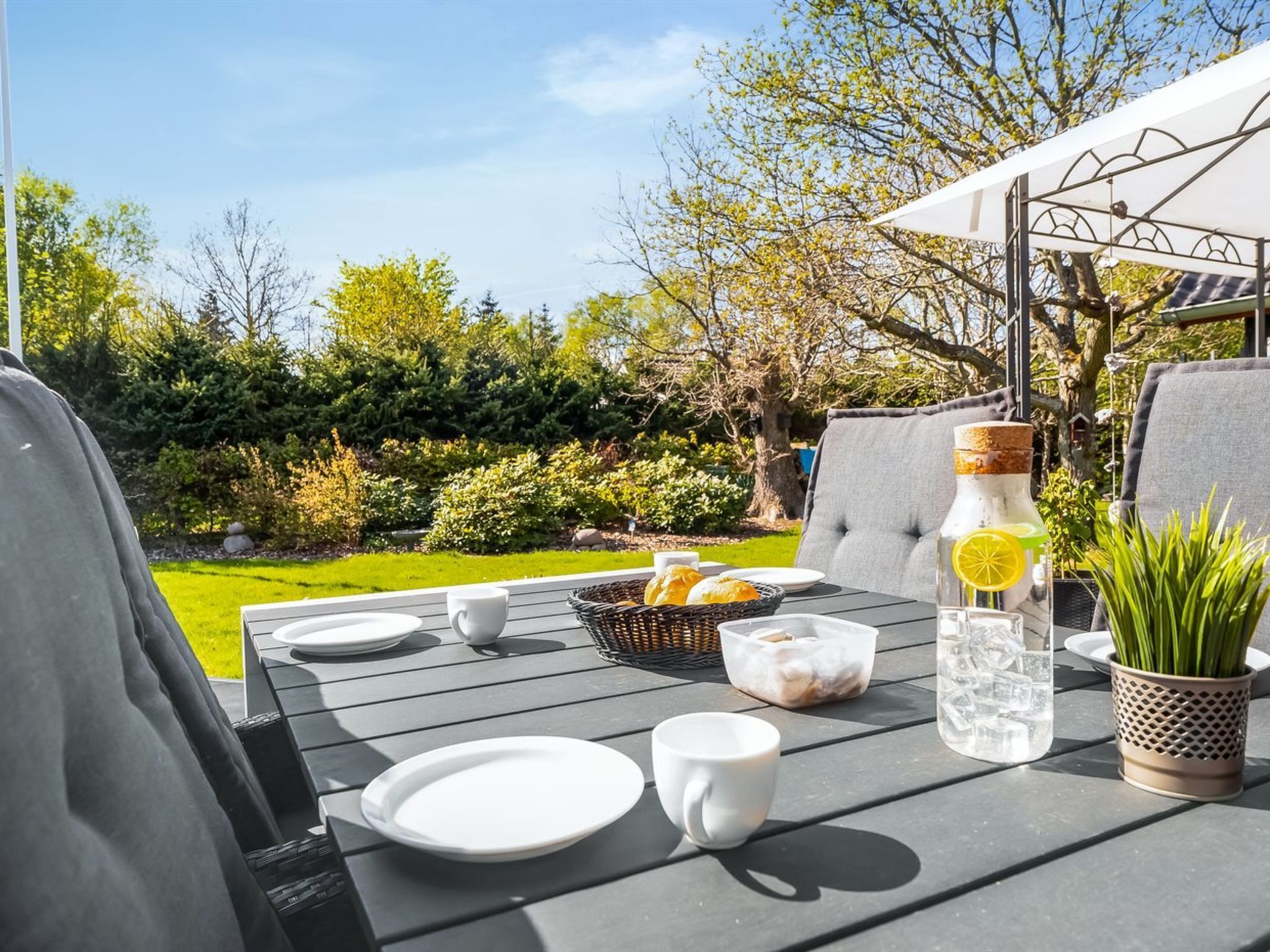 An outdoor dining area with a table set for two, featuring a basket of bread, a bottle of water, and a potted plant.