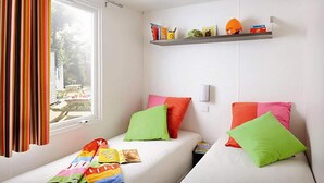 Een kamer met twee bedden, kleurrijke kussens, een raam met gordijnen en een plank met diverse spullen.