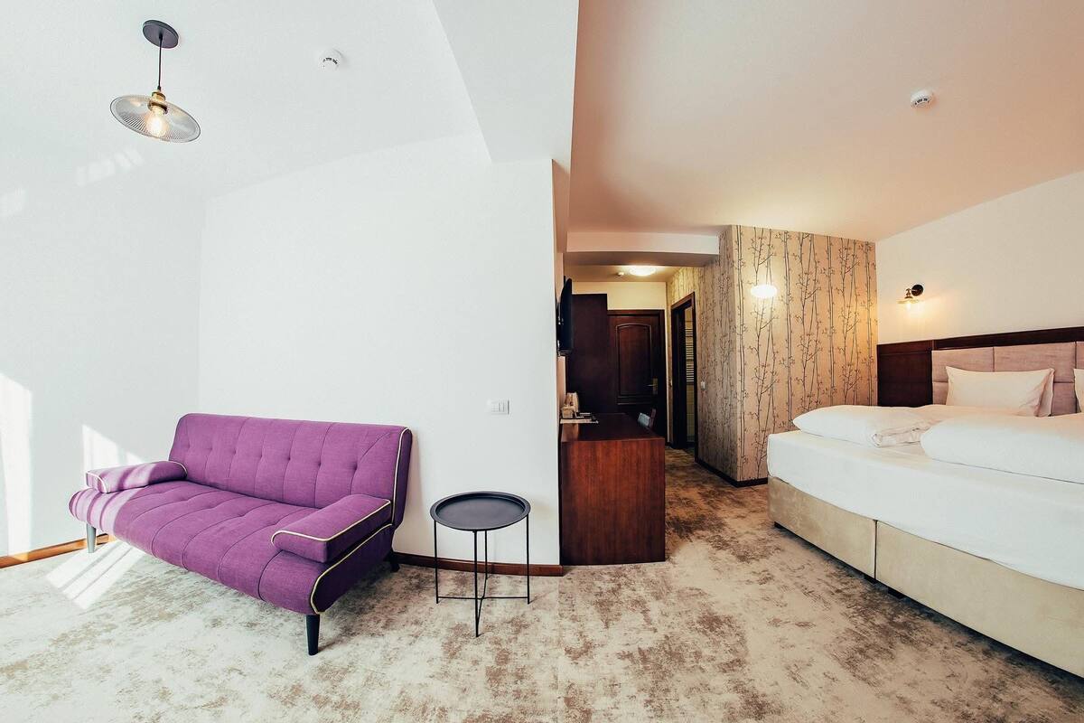 A hotel room with a purple sofa, a small round table, a bed, and a wooden desk.