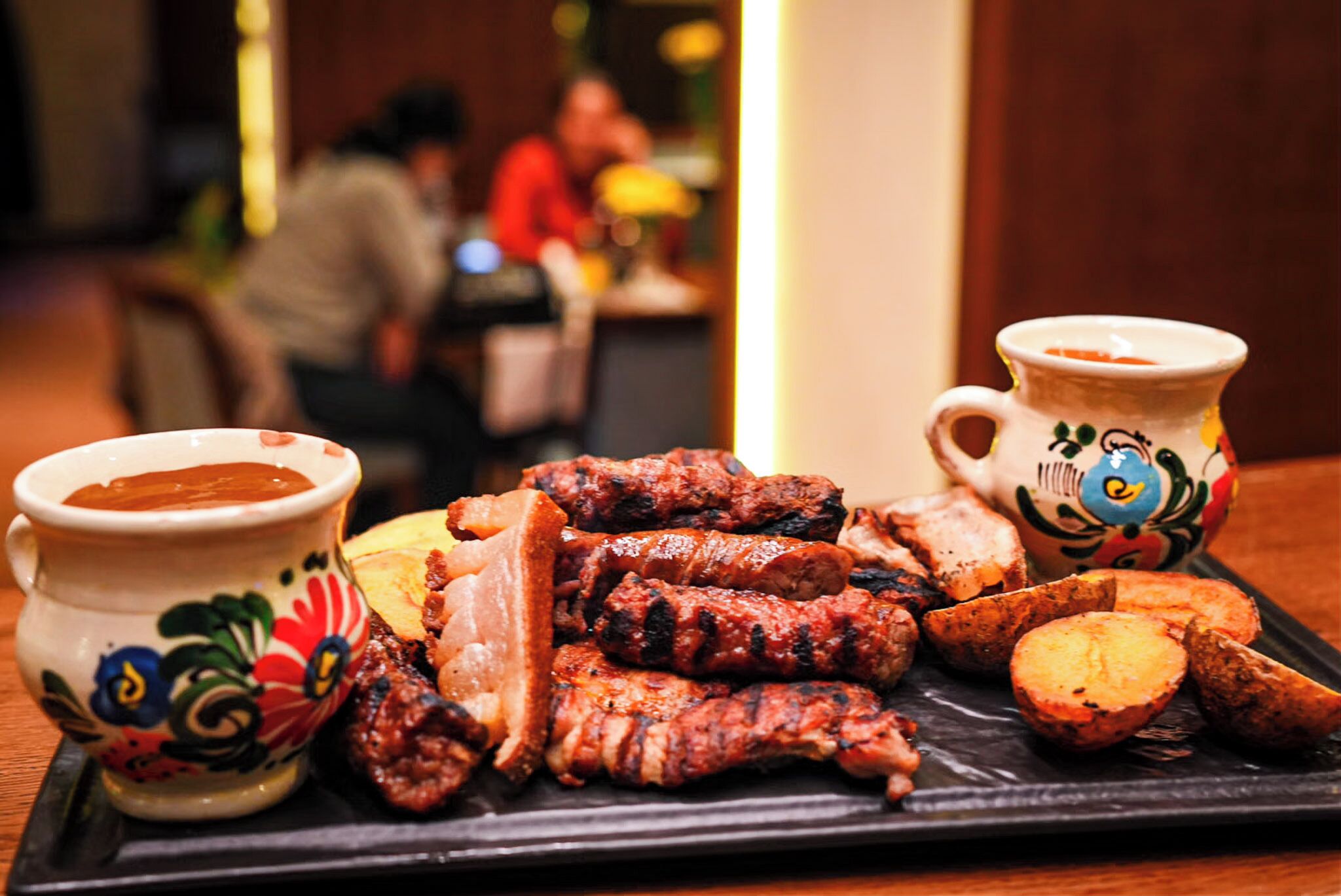 Una bandeja con carnes a la parrilla y papas, junto a dos vasos decorativos sobre una mesa.