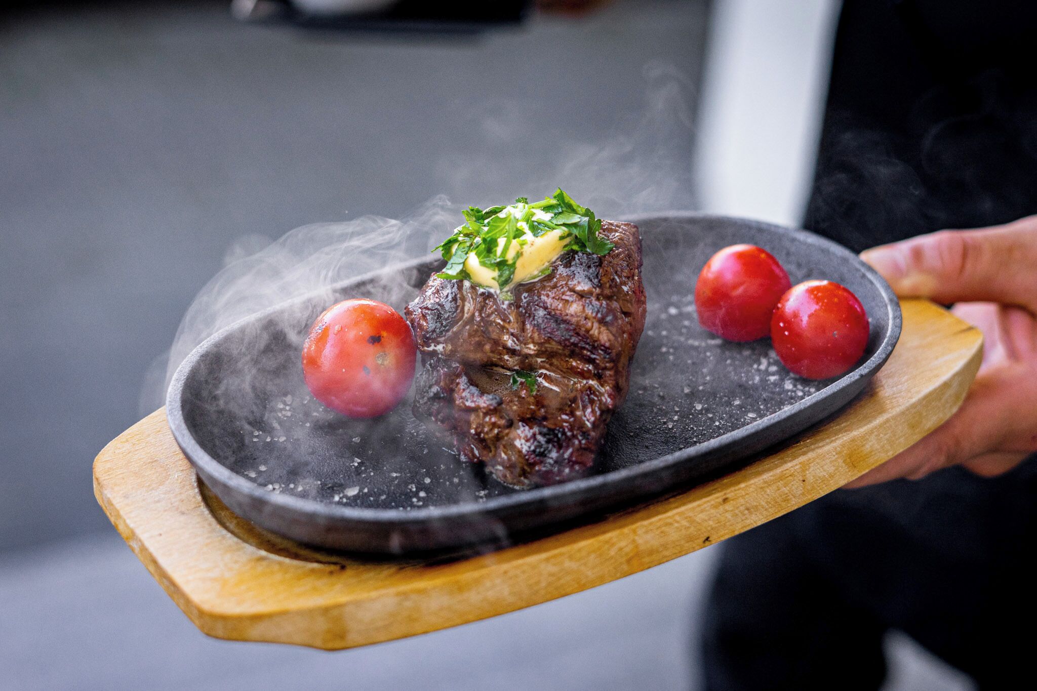 A steak with garnish and tomatoes on a black plate, held by a person.