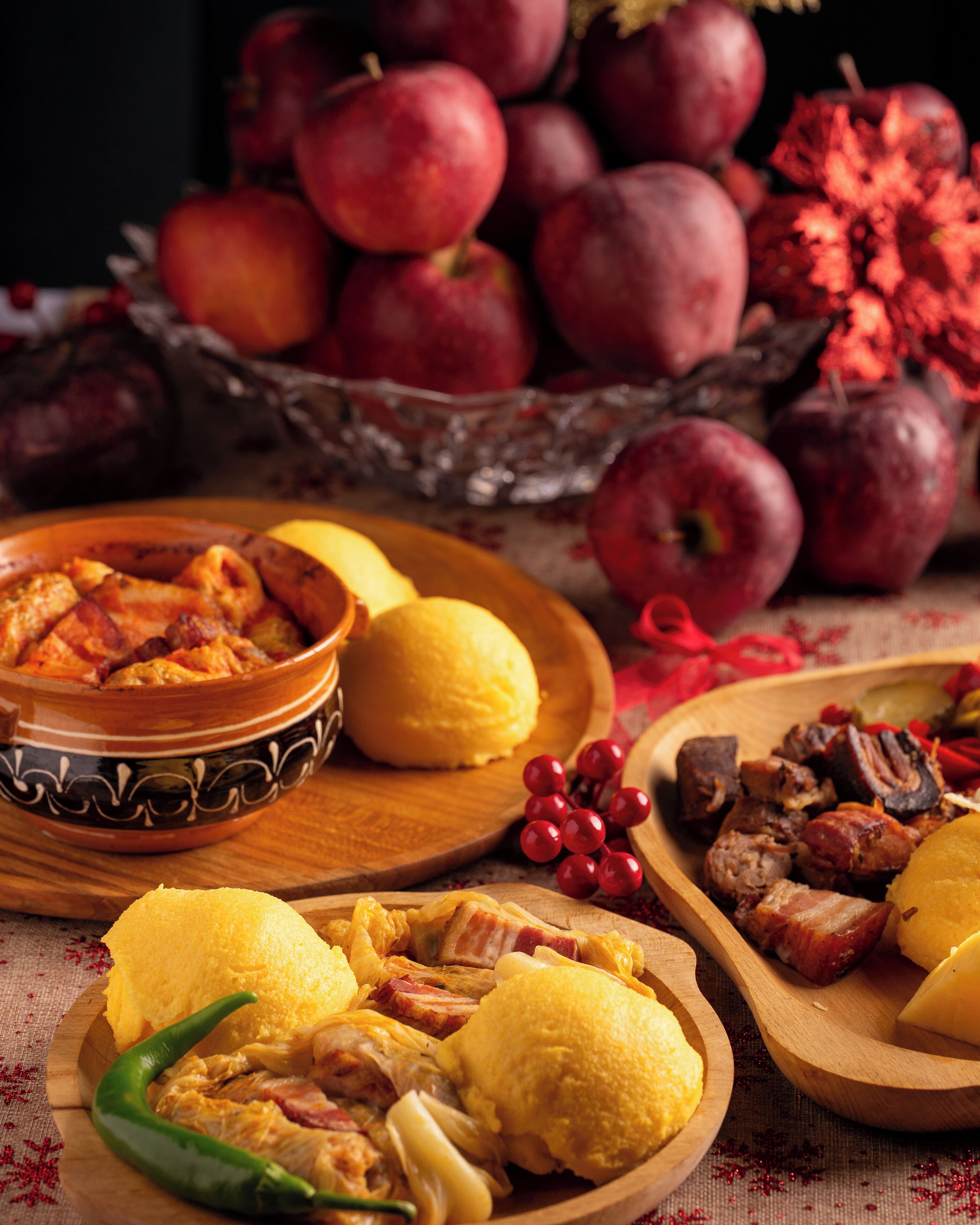 A wooden table with a variety of dishes including a bowl of food, a plate with meat and potatoes, and a bowl of yellow food. There are also red fruits and a green chili pepper visible.