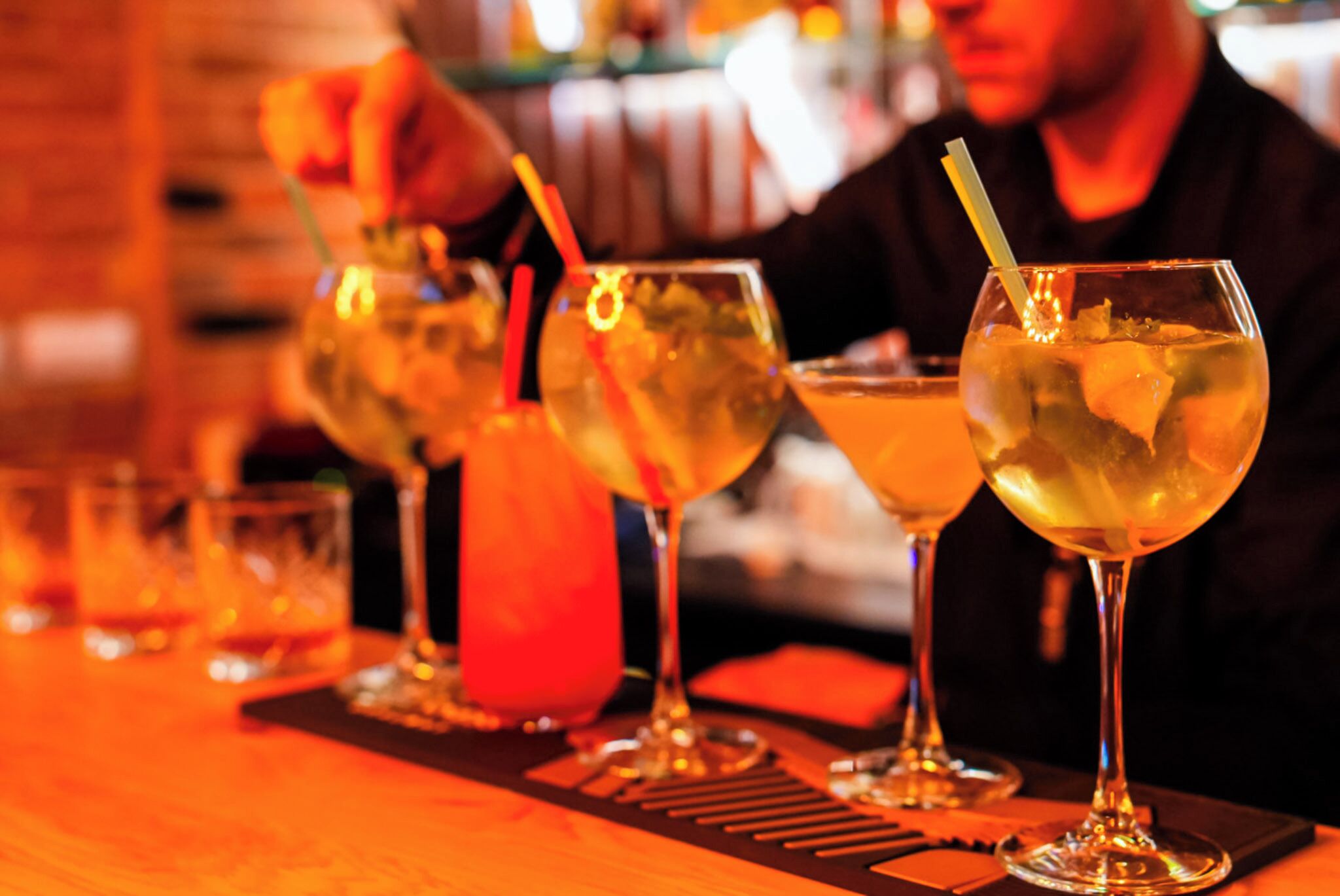 A bar setting with multiple glasses of beverages, a bartender, and a wooden bar counter.