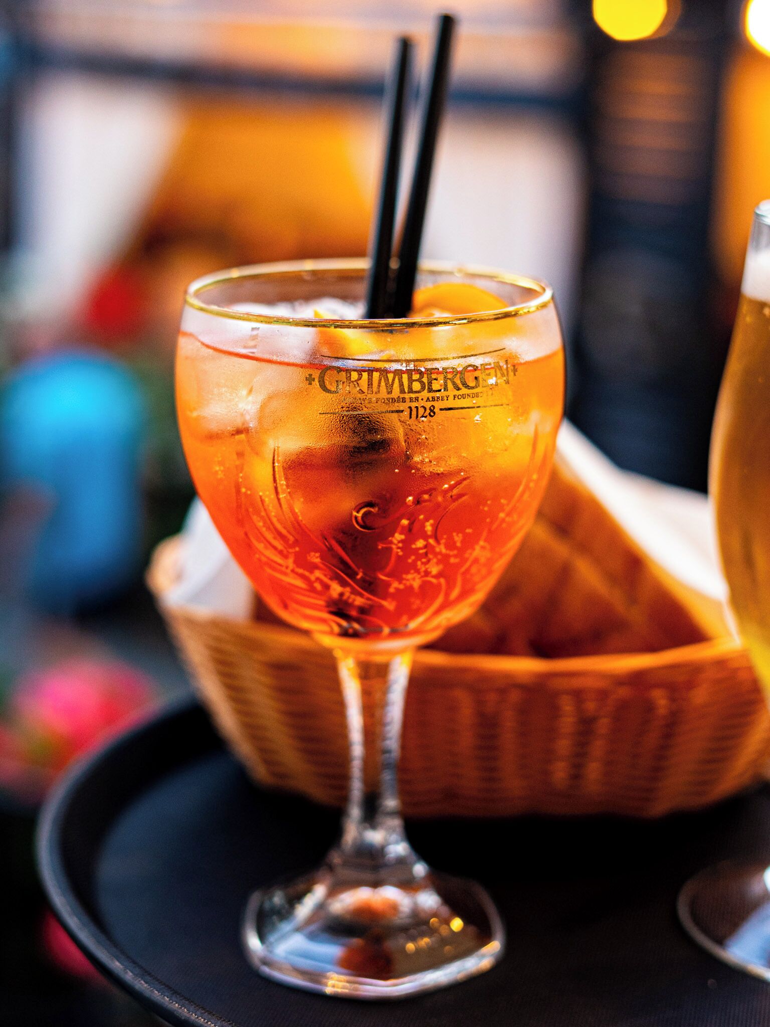 A glass of amber-colored beverage with a straw, placed on a black tray.
