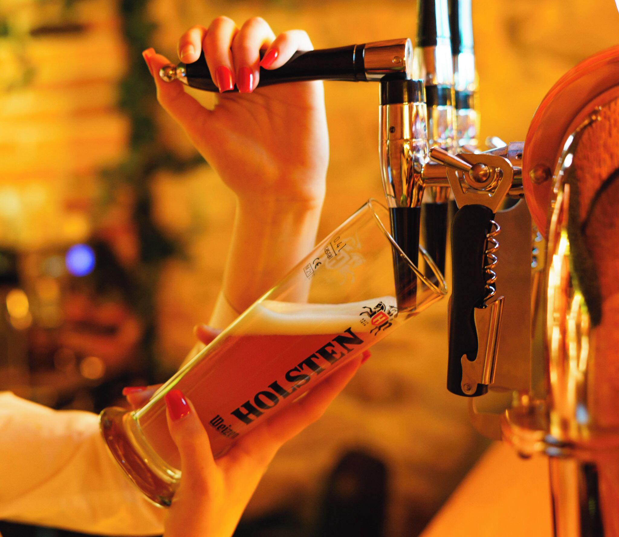 En hånd holder et glas med 'HOLSTEN' skrevet på, placeret under en fadølshaner.